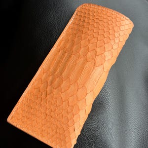 Pu&ograve; includere: Custodia per occhiali in pelle arancione con motivo a scaglie. La custodia rettangolare ha bordi arrotondati e cuciture bianche. La custodia &egrave; realizzata in un materiale strutturato.