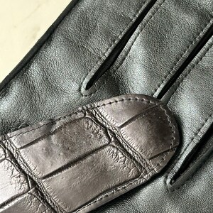 Pode incluir: Close-up de um par de luvas de couro pretas. As luvas apresentam uma tira texturizada em estilo crocodilo castanho escuro nas costas da m&atilde;o. O couro parece liso e flex&iacute;vel, com detalhes de costura vis&iacute;veis.