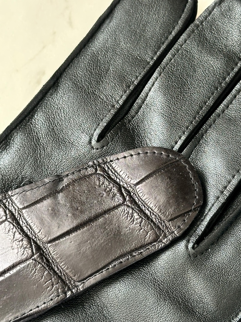 Pode incluir: Close-up de um par de luvas de couro pretas. As luvas apresentam uma tira texturizada em estilo crocodilo castanho escuro nas costas da m&atilde;o. O couro parece liso e flex&iacute;vel, com detalhes de costura vis&iacute;veis.