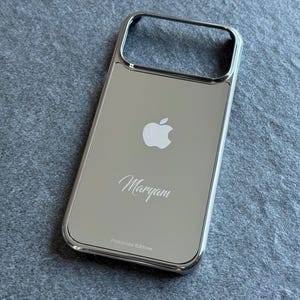 Pode incluir: Uma capa de telemóvel prateada e refletora com um acabamento metálico polido. A parte de trás apresenta um logótipo da Apple, o nome Maryam em letra cursiva e o texto Platinum Edition impresso na parte inferior. A capa tem um recorte retangular.