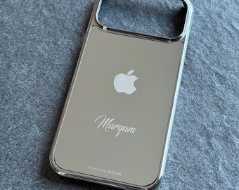 Op de afbeelding: Een zilverkleurige, reflecterende telefoonhoes met een gepolijste metalen afwerking. De achterkant is voorzien van een Apple-logo, de naam Maryam in cursief schrift en de tekst Platinum Edition onderaan. De hoes heeft een grote rechthoekige uitsparing.