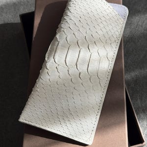 Op de afbeelding: Een lang, rechthoekig brillenkoker met een gestructureerde, gebroken witte slangenleren buitenkant met zichtbare overlappende schubben en gestikte randen, met een lichtgrijze binnenvoering bij de opening.