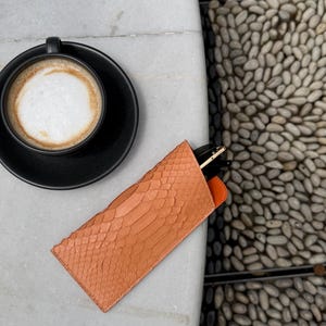 Pu&ograve; includere: Un astuccio per occhiali color pesca con texture di pelle di serpente e interno arancione poggia su una superficie di marmo bianco. Una tazza e un piattino da caff&egrave; neri sono sullo sfondo. L'astuccio contiene un paio di occhiali.