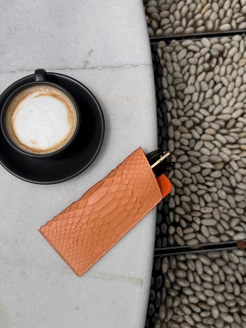 Pu&ograve; includere: Un astuccio per occhiali color pesca con texture di pelle di serpente e interno arancione poggia su una superficie di marmo bianco. Una tazza e un piattino da caff&egrave; neri sono sullo sfondo. L'astuccio contiene un paio di occhiali.