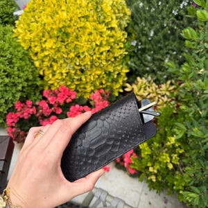 Pu&ograve; includere: Un astuccio nero con texture a pelle di serpente contiene un paio di occhiali da sole. L'astuccio &egrave; mostrato davanti a un giardino lussureggiante con fiori rosa e fogliame verde e giallo.