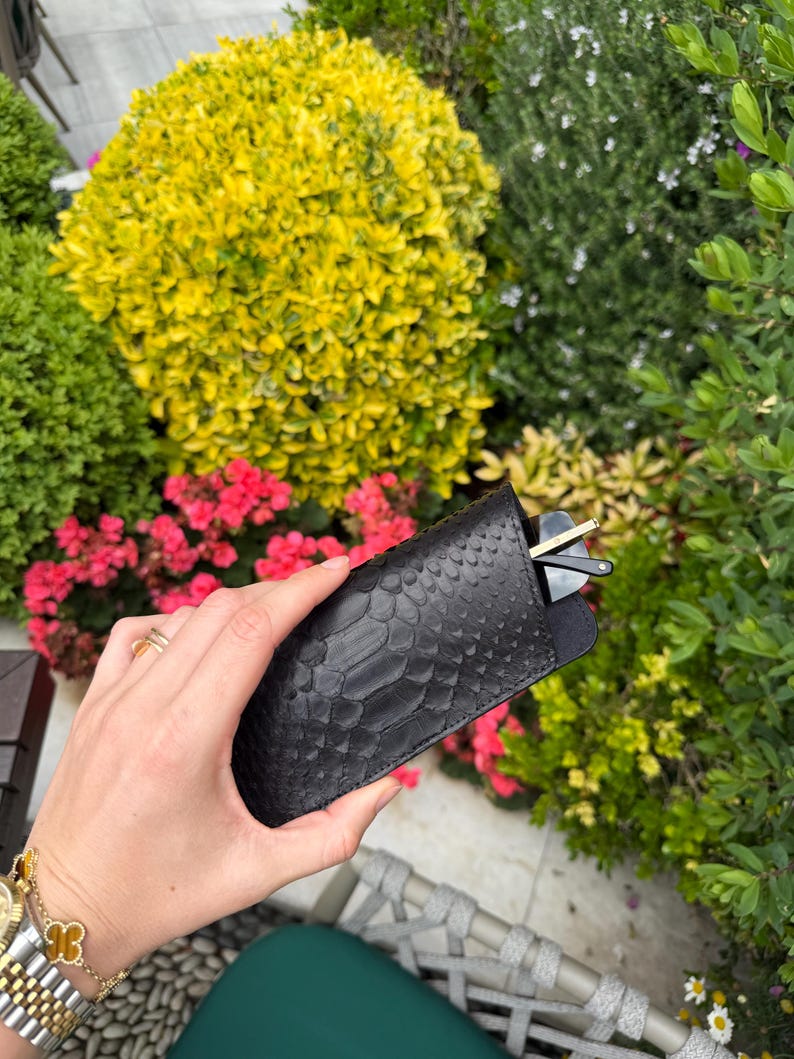 Pu&ograve; includere: Un astuccio nero con texture a pelle di serpente contiene un paio di occhiali da sole. L'astuccio &egrave; mostrato davanti a un giardino lussureggiante con fiori rosa e fogliame verde e giallo.