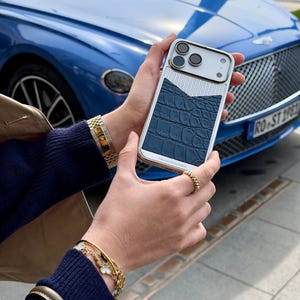 Pode incluir: Uma capa para smartphone com moldura metálica prateada, secção superior com riscas brancas e painel inferior em pele sintética azul escura com textura de crocodilo. Inclui um recorte para a câmara e o texto Platinum Drive Edition na base.