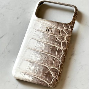 May include: A white and brown phone case with a crocodile skin pattern. The case has the initials "IG" in the upper center. The case is designed to fit a smartphone and has openings for the camera and buttons.