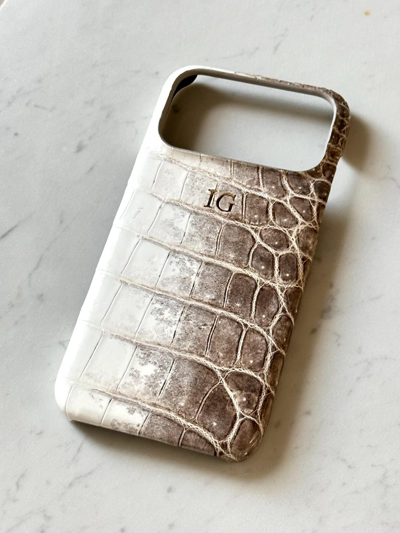 May include: A white and brown phone case with a crocodile skin pattern. The case has the initials "IG" in the upper center. The case is designed to fit a smartphone and has openings for the camera and buttons.