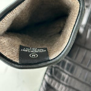 Pode incluir: Luvas de couro pretas com um padr&atilde;o de crocodilo texturizado. O interior &eacute; forrado com um material macio, castanho claro. Uma etiqueta dentro da luva indica que &eacute; 100% couro e tamanho M.