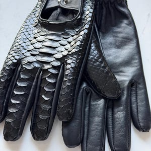 Könnte beinhalten: Ein Paar schwarze Lederhandschuhe. Einer der Handschuhe hat ein Schlangenmuster auf der Oberseite, der andere ist glatt. Die Handschuhe haben einen Druckknopfverschluss, für das Fahren oder die Mode.