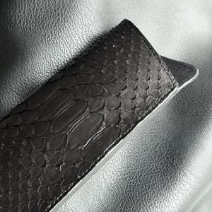 Pu&ograve; includere: Un astuccio per occhiali in pelle grigio scuro strutturata con un motivo a pelle di serpente. L'astuccio &egrave; aperto, rivelando un interno grigio chiaro. Lo sfondo &egrave; una superficie strutturata grigio scuro.