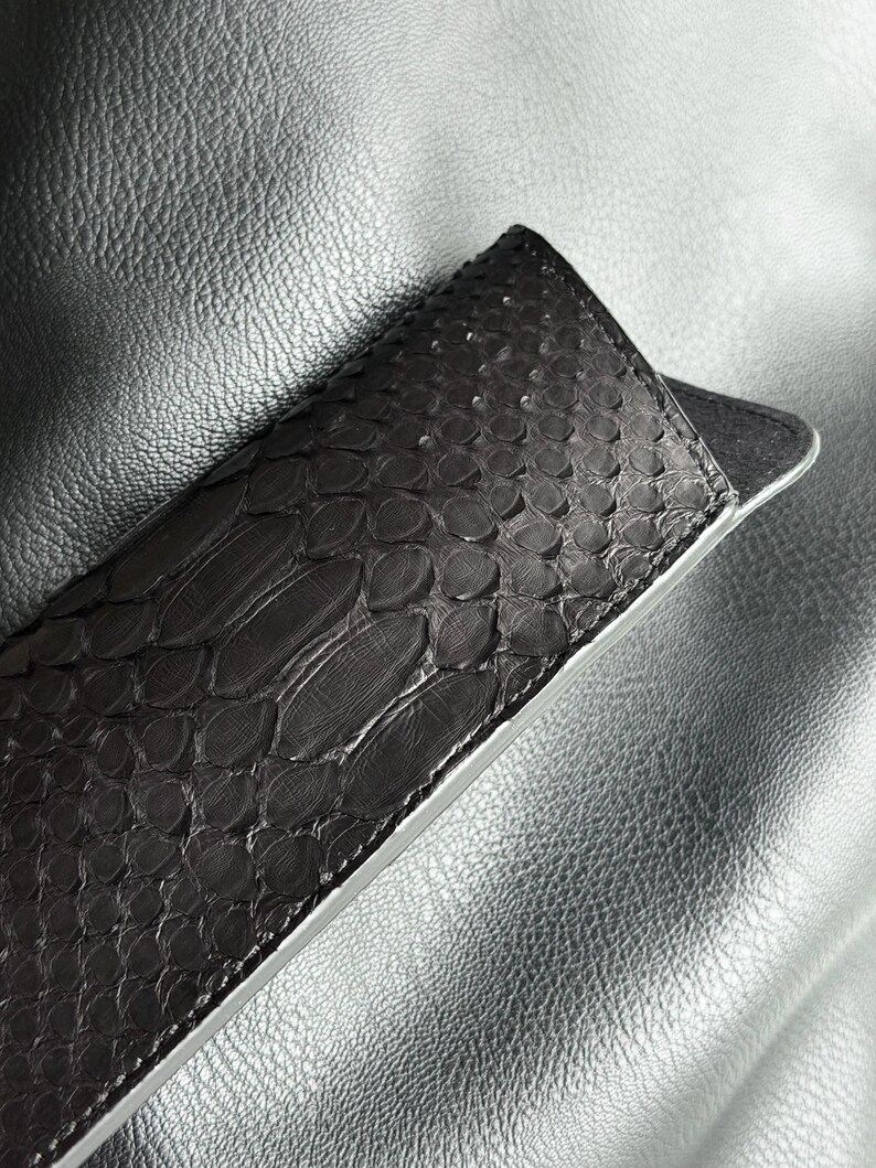 Pu&ograve; includere: Un astuccio per occhiali in pelle grigio scuro strutturata con un motivo a pelle di serpente. L'astuccio &egrave; aperto, rivelando un interno grigio chiaro. Lo sfondo &egrave; una superficie strutturata grigio scuro.
