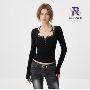 May include: A black long-sleeve top featuring a square neckline with black lace trim, a button-down placket, and two small black bows at the shoulders. The top is paired with dark gray denim jeans featuring a black belt with a silver buckle.