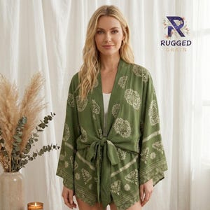 May include: An olive green kimono-style wrap top featuring a cream-colored botanical and geometric print. The garment has wide sleeves, an open front, and a matching fabric tie at the waist. The logo RUGGED GRAIN is visible in the upper right corner.