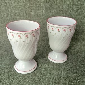 Può includere: Due calici in ceramica bianca con un motivo a spirale e bordo rosso. I calici hanno un motivo floreale intorno alla parte superiore e un accento rosso sulla base. I calici sono su uno sfondo verde.