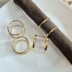 Könnte beinhalten: Ein weißer Teller mit einer gerollten braunen Serviette und goldenen und transparenten Serviettenringen. Die Serviettenringe haben eine transparente Mitte mit goldenem Rand. Zwei goldene Ringe liegen auf dem Teller.