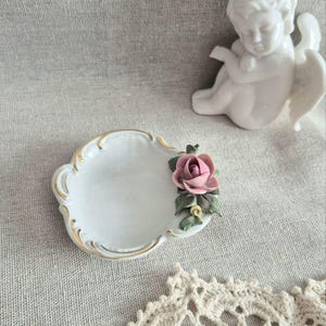 Könnte beinhalten: Eine weiße Porzellanschale mit Goldrand und einer modellierten rosa Rose mit grünen Blättern an der Seite, begleitet von einer kleinen weißen Porzellanfigur eines Cherubs mit Flügeln, platziert neben einem Stück weißer Häkelspitze.
