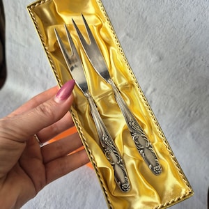 Pode incluir: Dois garfos de servir prateados com cabos ornamentados, apresentados numa caixa dourada com forro de cetim. Os garfos têm três dentes e são provavelmente vintage, adequados para servir aperitivos ou sobremesas.