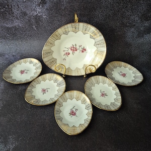 Könnte beinhalten: Ein Vintage-Servierset aus Porzellan mit einem großen, gewellten Teller mit goldenem Griff und sechs kleineren, ovalen Tellern. Jedes Stück hat eine cremefarbene Mitte mit floralem Design und goldenem Rand. Das Set ist auf einer dunklen Oberfläche angeordnet.