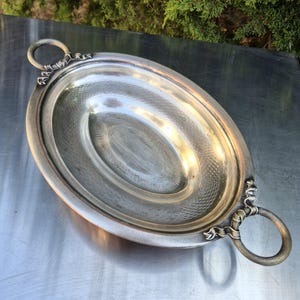 Pode incluir: Travessa oval de prata com textura martelada e alças em anel. A travessa tem uma borda elevada e detalhes decorativos perto das alças. Uma peça clássica para servir alimentos em ocasiões especiais.