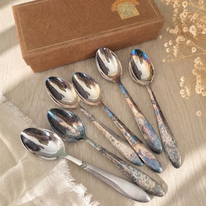 Puede incluir: Un juego de cinco cucharas de metal plateado con diversos grados de deslustre oscuro e iridiscente y un patrón de chevrón en el cuello del mango. Las cucharas están dispuestas en abanico junto a una caja rectangular marrón con una etiqueta dorada.