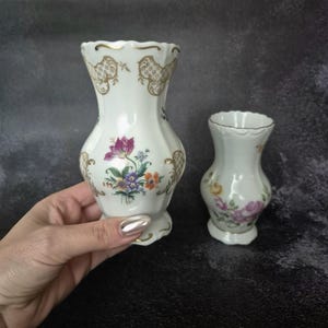 May include: Two white porcelain vases with floral designs. The larger vase features a purple flower with orange and blue accents, and gold filigree. The smaller vase has a similar design. Both have scalloped rims.