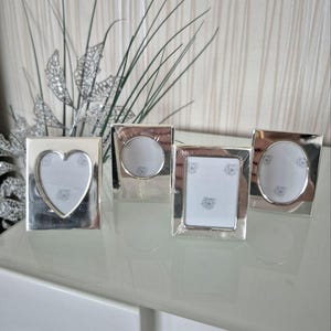 Peut inclure: Quatre cadres photo argentés de formes variées : un cœur, un cercle, un carré et un ovale. Chaque cadre contient un insert blanc avec un motif floral subtil. Les cadres sont disposés sur une surface réfléchissante.