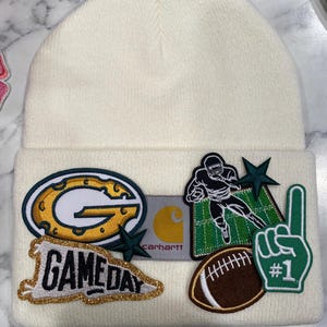 May include: Cream-colored beanie with multiple embroidered patches. Patches include a football, a football player, a foam finger, and the words "Game Day". The Carhartt logo is also visible.
