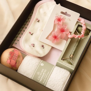 May include: A gift box containing a variety of self-care items. Includes a pair of floral socks, a jade roller, a wooden ball with text, a hair accessory with flower accents, a notepad, and two white face cloths.