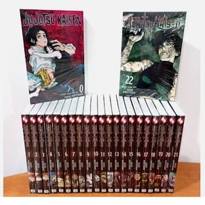 Puede incluir: Una colección de volúmenes de manga Jujutsu Kaisen. La imagen muestra los volúmenes 0 y 22 sobre una fila de volúmenes del 1 al 21. Las portadas presentan ilustraciones de personajes y el título de la serie. Los lomos son rojos con texto blanco. Los libros están sobre una superficie de madera.