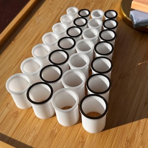 Puede incluir: Una colección de objetos cilíndricos blancos con bordes negros, dispuestos sobre una superficie de madera marrón claro. Los objetos están espaciados uniformemente y parecen ser pequeños recipientes o componentes, posiblemente para manualidades u otros artículos pequeños.