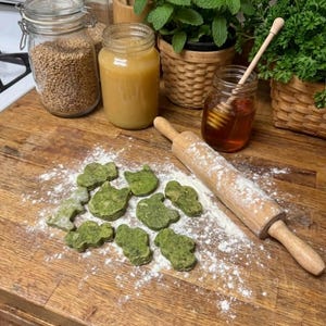 May include: A wooden rolling pin rests on a wooden surface dusted with flour, next to green, cat-shaped cookies. Jars of honey, grains, and herbs are in the background, suggesting a baking or cooking theme.