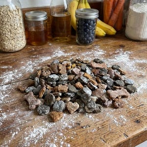 Pu&ograve; includere: Un mucchio di biscotti per cani di varie forme e colori, tra cui ossa e cuori, su una superficie di legno rustico spolverata di farina. Vasi di avena, miele, mirtilli e altri ingredienti sono sullo sfondo.
