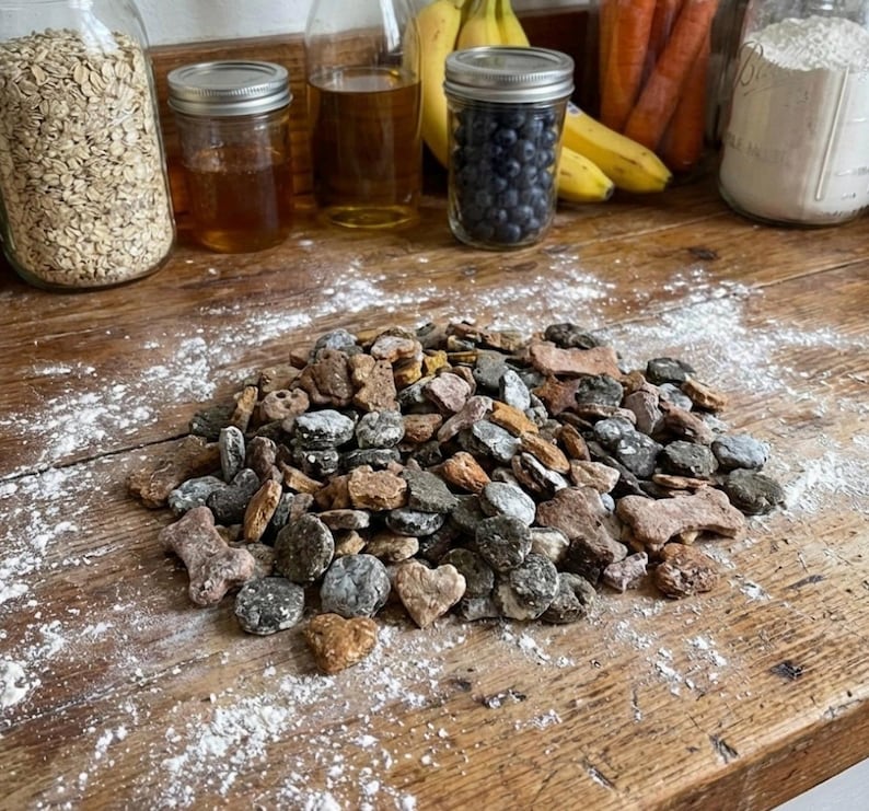 Pu&ograve; includere: Un mucchio di biscotti per cani di varie forme e colori, tra cui ossa e cuori, su una superficie di legno rustico spolverata di farina. Vasi di avena, miele, mirtilli e altri ingredienti sono sullo sfondo.