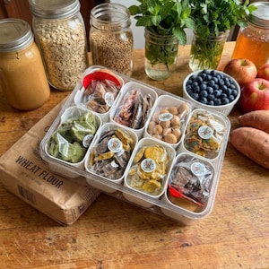 Puede incluir: Una mesa de madera exhibe una variedad de alimentos. Un recipiente de pl&aacute;stico transparente contiene varios peque&ntilde;os dulces envasados individualmente. Se ven frascos de avena, trigo y mantequilla de man&iacute;, junto con menta fresca, ar&aacute;ndanos, manzanas y batatas.