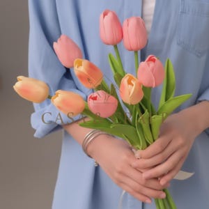 以下が含まれることがあります： ピンクとピーチの色合いの人工チューリップのブーケ。緑色の茎と葉が付いています。白いリボンでまとめられています。「CASA」の文字が画像に表示されています。