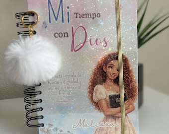 May include: A spiral-bound notebook with a holographic finish featuring the text Mi Tiempo con Dios and a girl holding a book. The cover includes the Spanish text está vestida de fuerza y dignidad y sonríe sin temor al futuro. A white fluffy pom-pom keychain is attached to the black spiral binding.