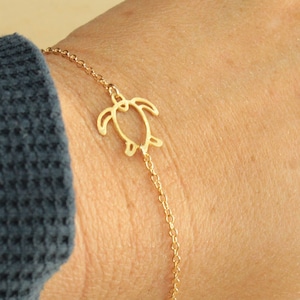 Peut inclure: Un bracelet en chaîne dorée avec un délicat pendentif en forme de tortue. Le bracelet est porté au poignet.
