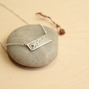 May include: A silver necklace with a rectangular pendant featuring a detailed dandelion design with seeds blowing in the wind.