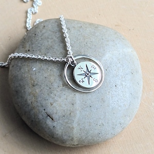 May include: A silver necklace featuring a circular pendant with a compass rose design. The pendant is suspended from a delicate silver chain. The compass rose is engraved with the letters N, S, E, and W, representing the cardinal directions.