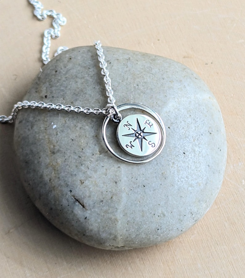 May include: A silver necklace featuring a circular pendant with a compass rose design. The pendant is suspended from a delicate silver chain. The compass rose is engraved with the letters N, S, E, and W, representing the cardinal directions.