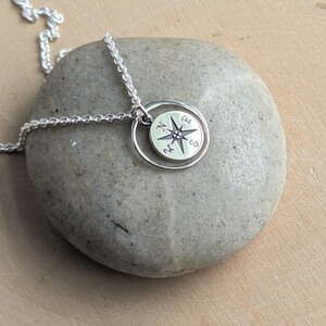 May include: A silver necklace featuring a circular pendant with a compass rose design. The pendant is suspended from a delicate silver chain and displays the cardinal directions N, E, S, and W engraved on its surface within a thin silver ring.