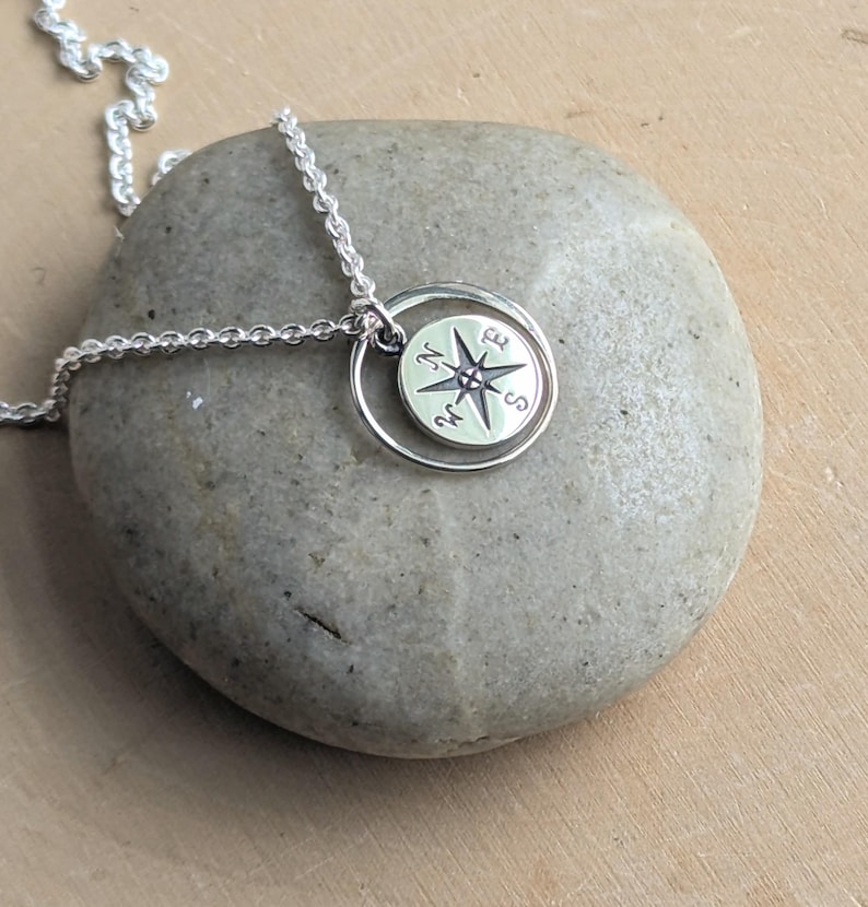 May include: A silver necklace featuring a circular pendant with a compass rose design. The pendant is suspended from a delicate silver chain and displays the cardinal directions N, E, S, and W engraved on its surface within a thin silver ring.