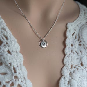 May include: A silver-toned necklace featuring a small, circular pendant with an engraved compass rose design. The pendant is suspended from a delicate, thin silver-toned chain. The compass rose includes the letters N, S, E, and W.