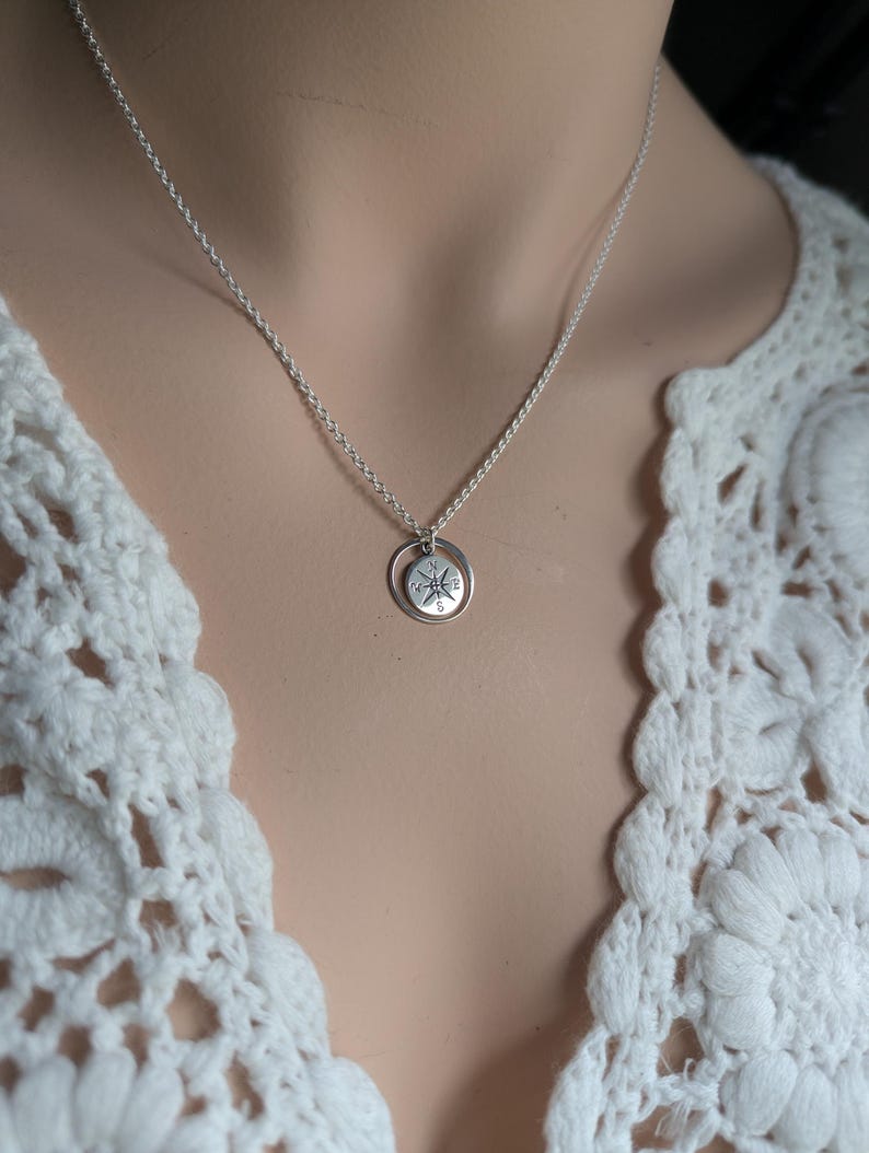 May include: A silver-toned necklace featuring a small, circular pendant with an engraved compass rose design. The pendant is suspended from a delicate, thin silver-toned chain. The compass rose includes the letters N, S, E, and W.