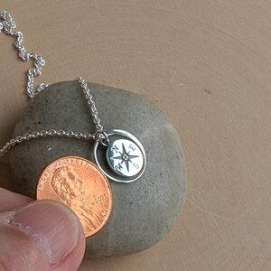 May include: A silver necklace featuring a small circular pendant with an engraved compass rose design showing the letters N, E, S, and W. The pendant is suspended from a delicate silver chain and is shown next to a copper penny for scale.