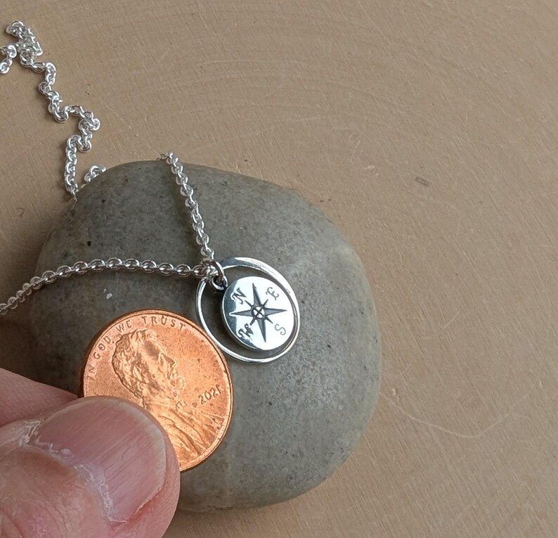 May include: A silver necklace featuring a small circular pendant with an engraved compass rose design showing the letters N, E, S, and W. The pendant is suspended from a delicate silver chain and is shown next to a copper penny for scale.