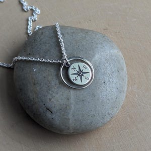 May include: A silver-coloured necklace featuring a circular pendant with a compass rose design. The pendant includes a small round disc engraved with the cardinal directions N, E, S, and W, suspended from a delicate silver-coloured chain link necklace.