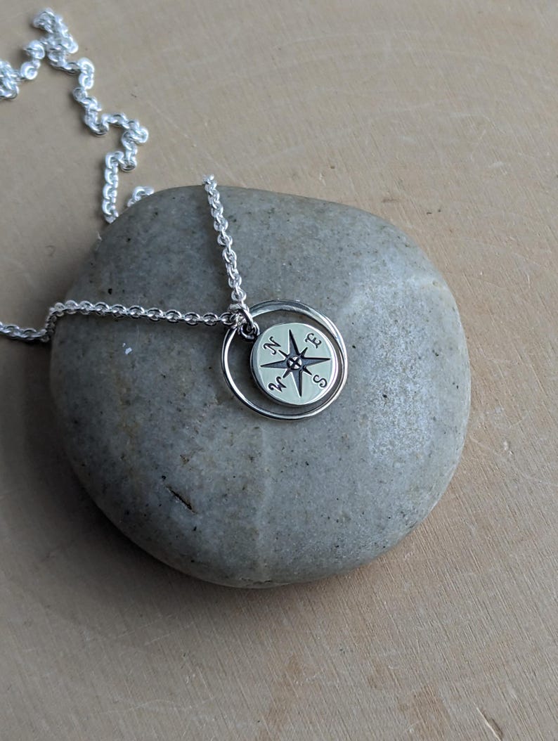 May include: A silver-coloured necklace featuring a circular pendant with a compass rose design. The pendant includes a small round disc engraved with the cardinal directions N, E, S, and W, suspended from a delicate silver-coloured chain link necklace.